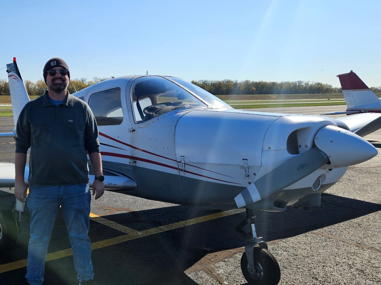 Dan smiling in a beanie and sunglasses, standing beside a white single-engine airplane on the ramp on a clear autumn day